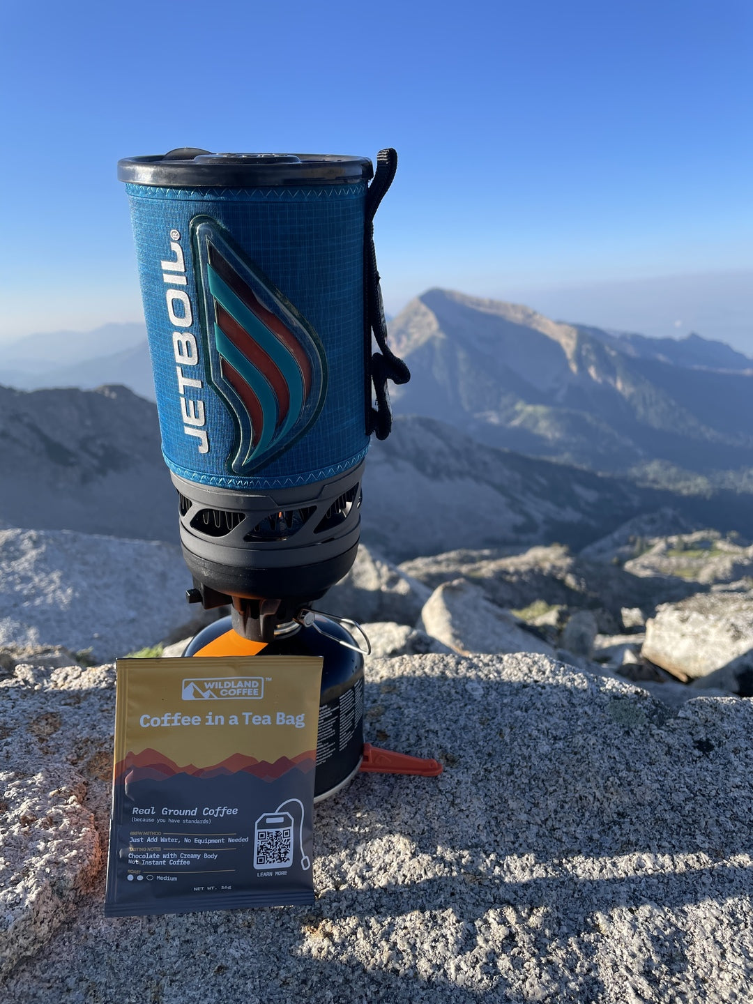 Picture of a Wildland Coffee pouch next to a Jetboil on the top of a mountain and overlooking other mountains. This coffee is perfect for on the go, hiking, backpacking, camping, and even just quick coffee at home.